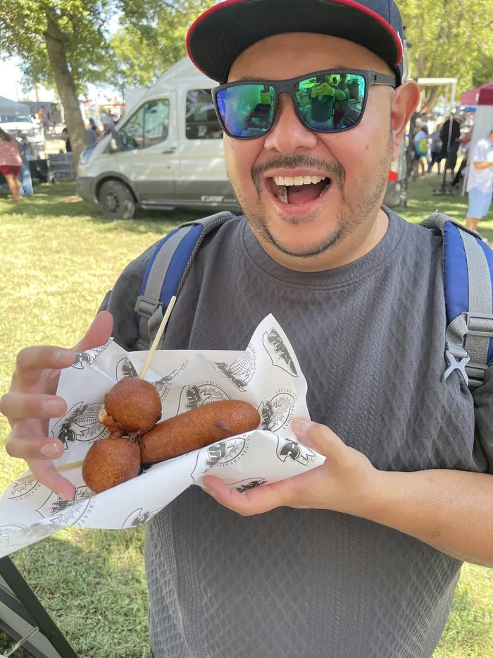 Sancho Combo: a smiling customer in sunglasses and a cap holds a corn dog and two cheeseballs on a stick, wrapped in Triple C paper, at a busy outdoor event.