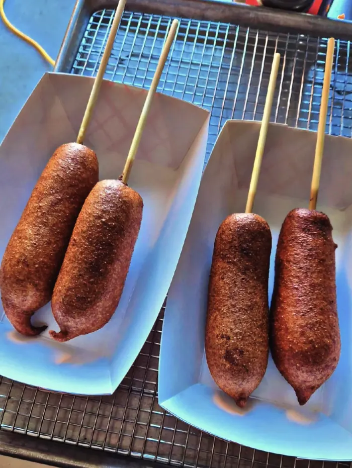 Four golden-brown corn dogs on wooden sticks in two paper food boats, resting on a cooling rack.
