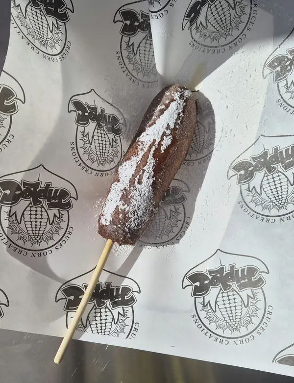 Breakfast Sausage on a Stick: a golden battered breakfast sausage on a stick, topped with powdered sugar and syrup, on white paper printed with the Triple C Cruces Corn Creations logo.